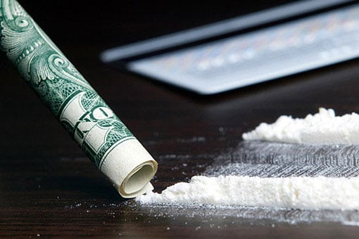 A razor blade lies next to two lines of cocaine and a rolled up dollar bill.